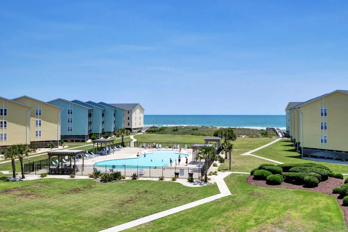 A cheerful outdoor scene featuring a large swimming pool set against the backdrop of the beach and the ocean. Surrounding buildings and well-maintained gardens frame the area, with guests enjoying the pool and sunbathing.