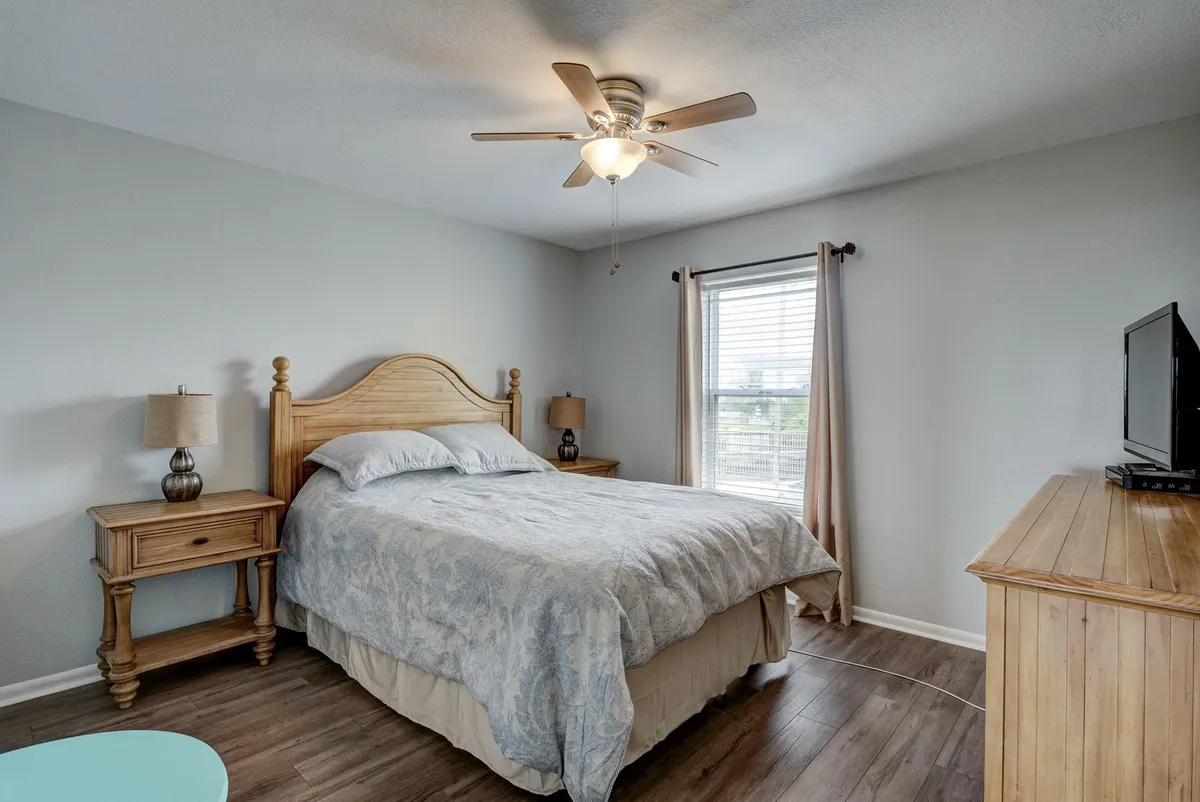 A serene bedroom featuring a plush queen bed with light blue bedding, flanked by wooden nightstands and illuminated by natural light from a curtained window. A ceiling fan and wall-mounted television add comfort and entertainment, making this space perfect for relaxation.