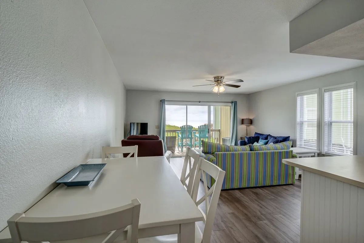 The scene showcases a dining table situated near a colorful couch, with a clear view through a sliding door to the outside. The setup is ideal for group meals, casual conversation, and enjoying sunsets or ocean vistas from the comfort of the condo.