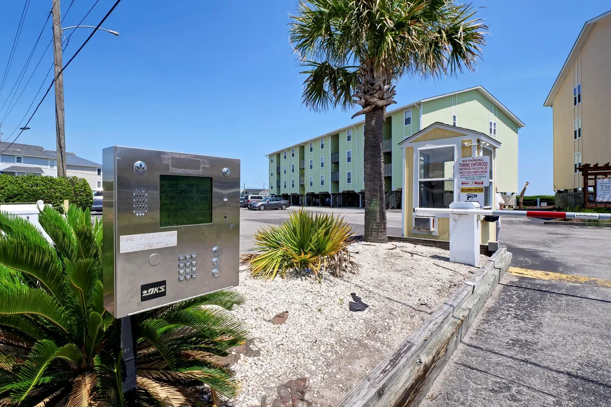 This exterior scene captures the welcoming entrance area to the property, with palm trees, a green building, and an entrance booth. The relaxed, friendly vibes set the stage for guests arriving for a special retreat, suggesting a hassle-free check-in and warm hospitality.
