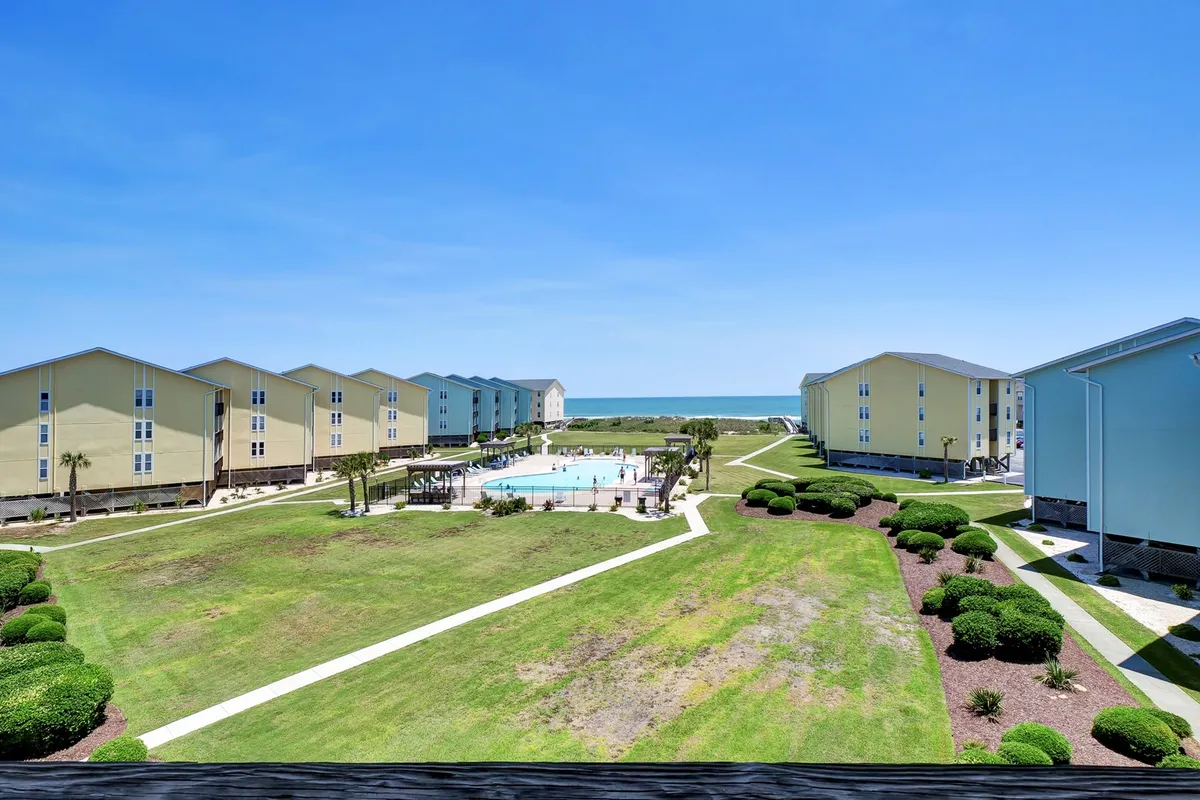 A wide view of the sparkling swimming pool surrounded by landscaped gardens, with the ocean visible in the distance and nearby resort buildings framing the scene. Lounge chairs and umbrellas are arranged around the pool, inviting guests to relax, swim, or enjoy the sunshine.