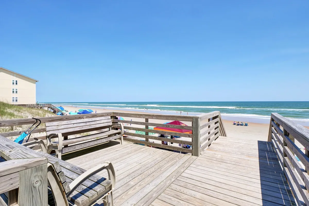 A wide view of a wooden deck overlooking the beach, with colorful beach chairs and umbrellas set up near the shoreline. The ocean is visible in the background, and the scene is filled with playful, inviting energy suitable for sunbathing, swimming, and family picnics.