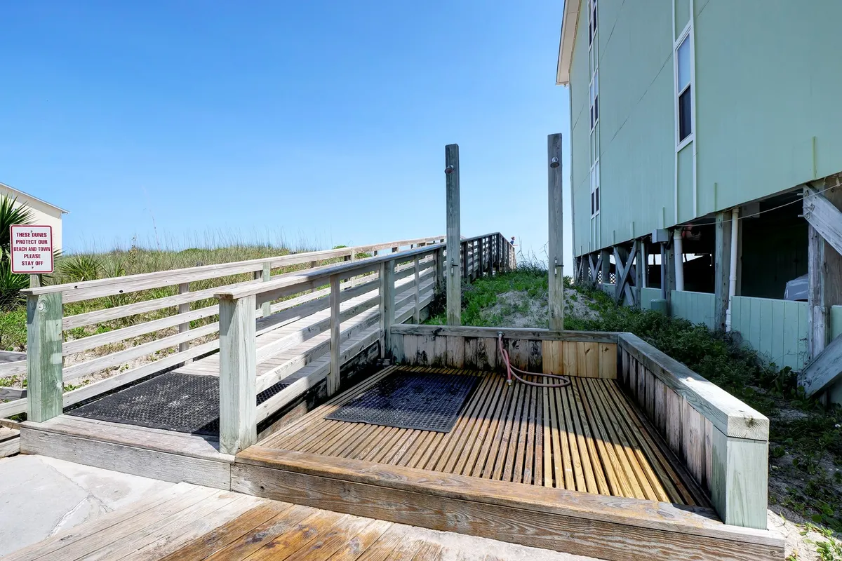 A wooden walkway leads through lush greenery to a shower area and onward toward the beach, providing a sense of transition between the property and the oceanfront. The space is serene and inviting, ideal for post-beach rinses or leisurely strolls to the water.