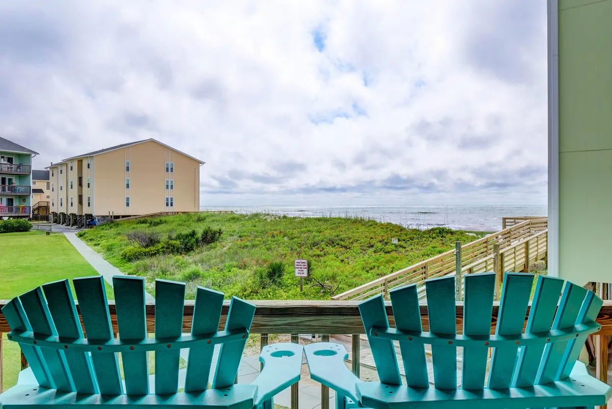 The hero image features two inviting turquoise Adirondack chairs positioned on a balcony, overlooking a sweeping ocean view with a lush green lawn below. The tranquil scene suggests moments of relaxation, where guests can watch dolphins in the distance or unwind with a coffee at sunrise or a glass of wine at sunset.