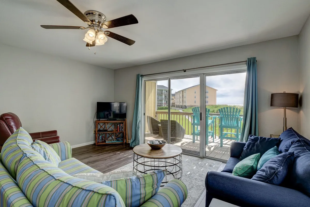 A comfortable, casual living area with a striped sofa, blue armchair, and a coffee table, all oriented toward large sliding glass doors leading to a balcony with an ocean view. The space hints at cozy movie nights, casual meals, and easy access to the outdoor deck for sunrise or sunset moments.