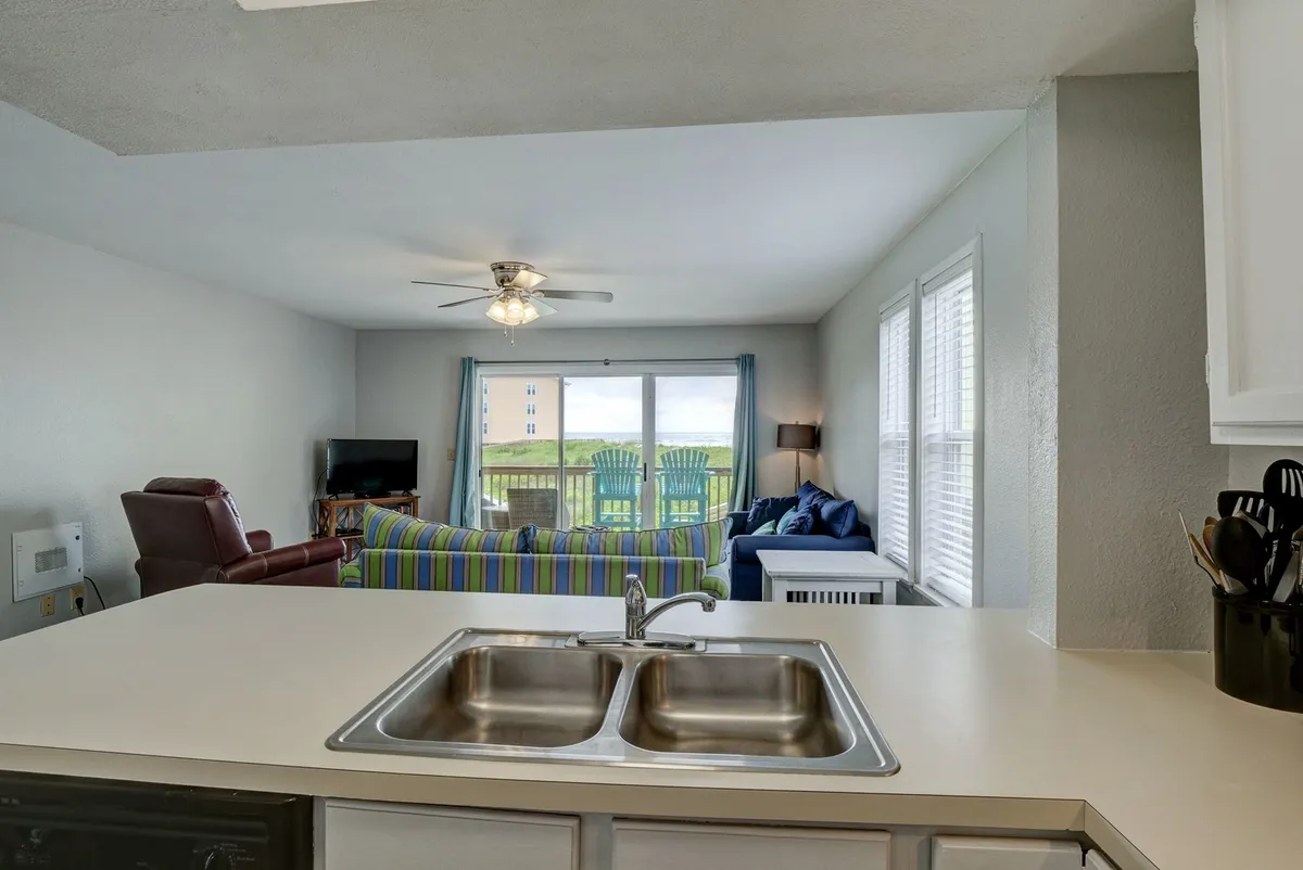 This image features a kitchen sink area that opens up to a living room with comfortable seating and a balcony view, providing a sense of openness and connection to the outdoors. The arrangement encourages group activities like cooking, entertaining, and relaxing, with the balcony inviting guests to gather and enjoy ocean views.