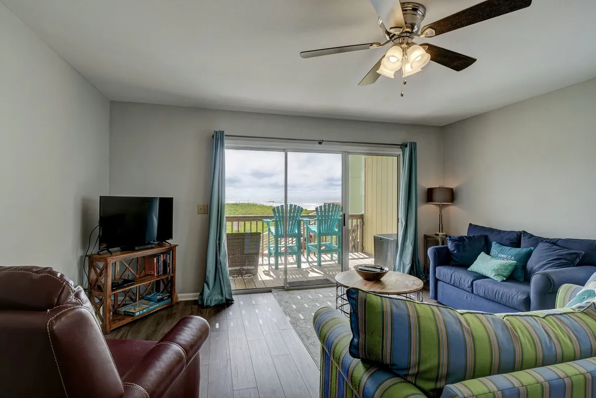 This image showcases a spacious, sunlit living area anchored by a large sliding glass door that opens to an oceanview deck. The inviting space is furnished with colorful couches and a wooden coffee table, with a TV as a focal point. The overall mood is cozy yet vibrant, suggesting a perfect setting for group relaxation or family gatherings while enjoying seaside views.